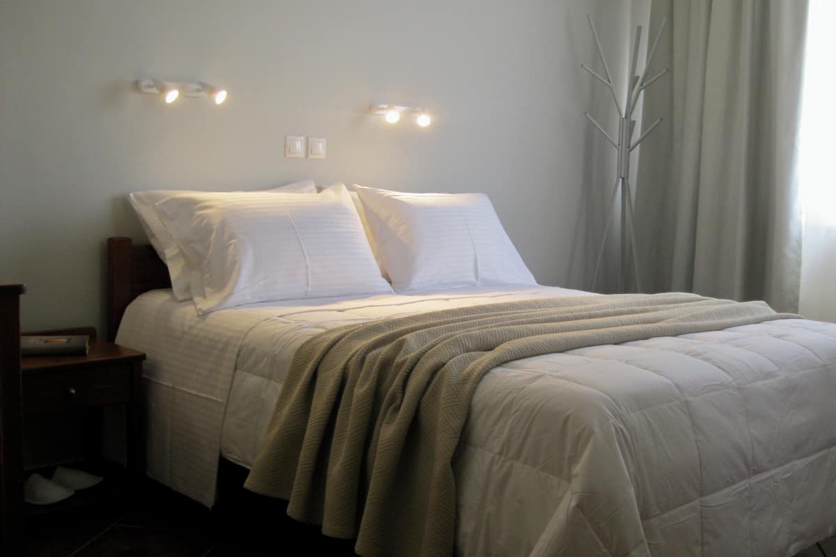 Un lit bien fait, avec une couette blanche et des oreillers, une table de chevet avec une lampe, et une fenêtre ornée de rideaux légers.