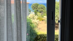 A view through a window with sheer curtains, revealing a garden with various plants and a distant view of the sea. - Iolkos Hotel Apartments (Chania)
