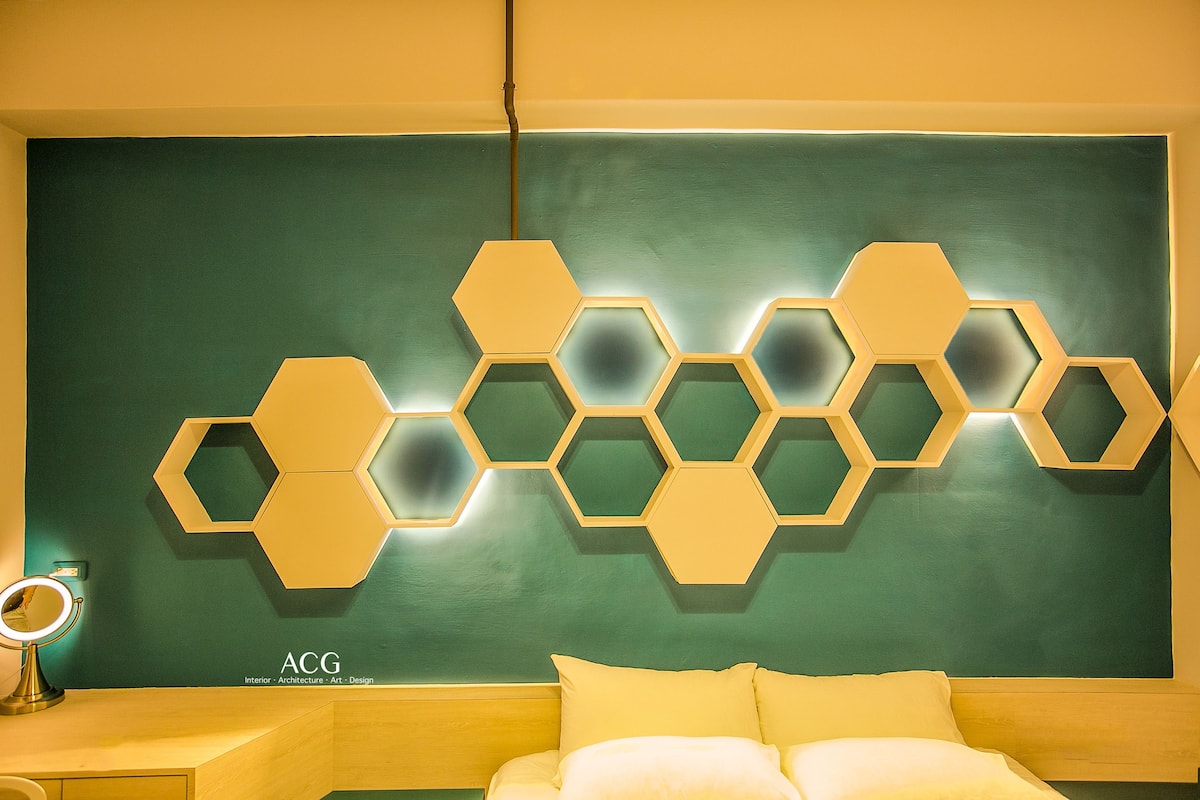 A room with a green wall, a wooden headboard, and a yellow bed. There is a decorative hexagonal light fixture on the wall.