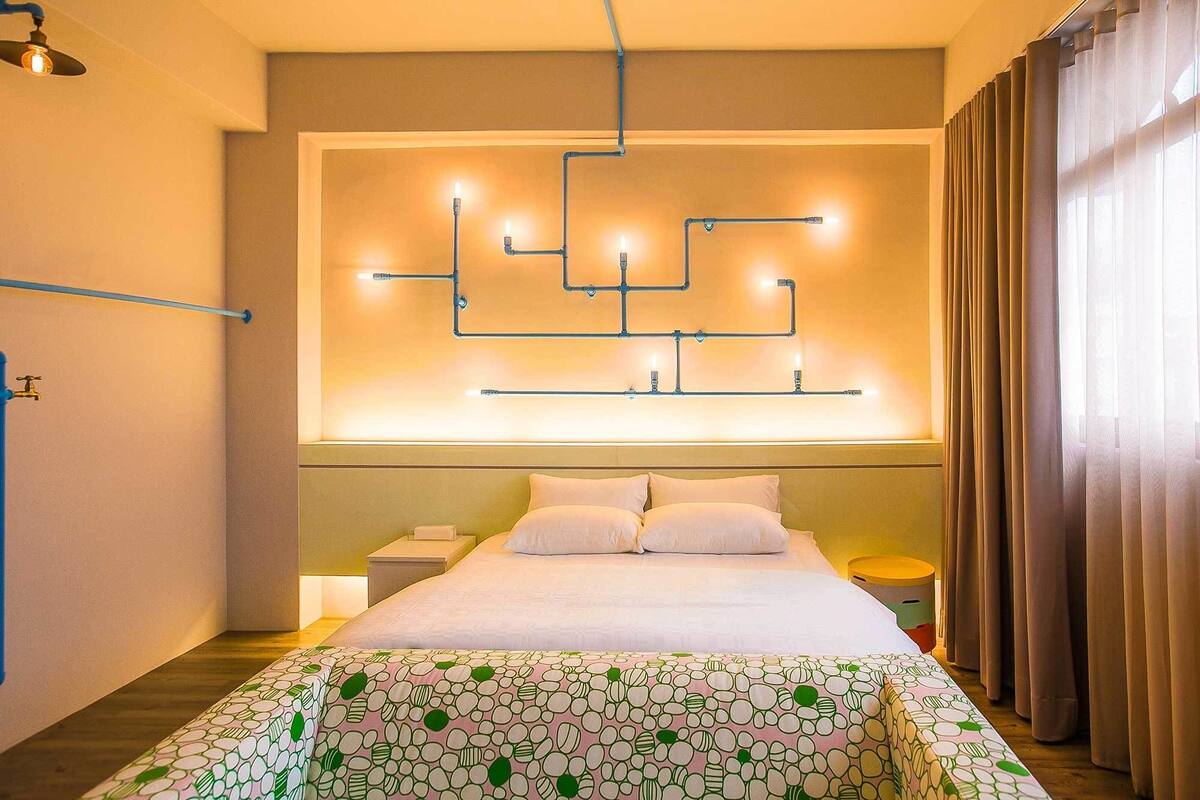 A modern hotel room with a bed, a headboard with integrated lighting, a bedside table, and a window with curtains.