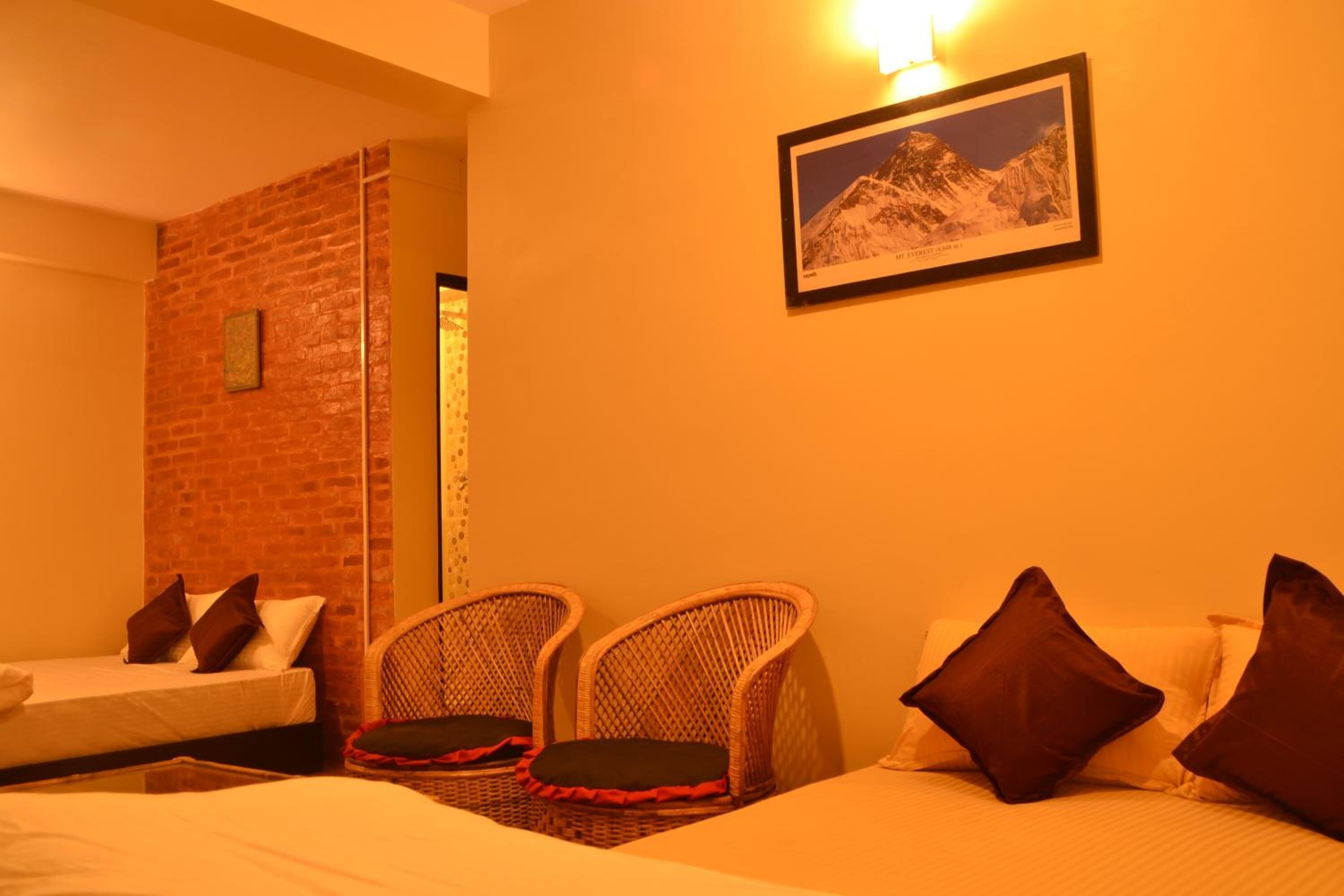 Quarto de hotel com parede de destaque em tijolo, cama com travesseiros, duas cadeiras de vime e um quadro pendurado na parede.