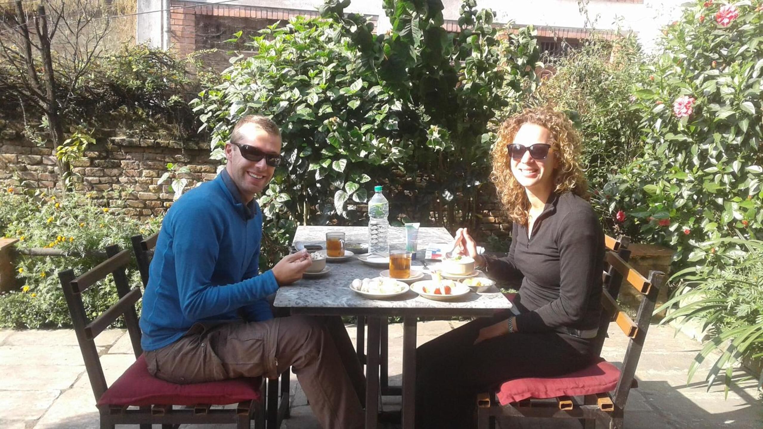 Dos personas sentadas en una mesa al aire libre, con comida y bebidas, rodeadas de follaje y un muro de ladrillo.