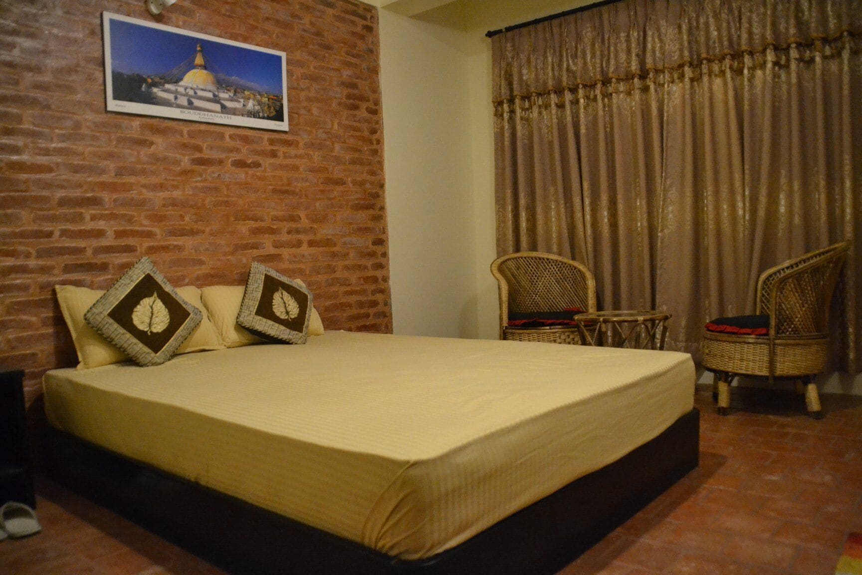 Een hotelkamer met een bakstenen muur, een bed met een beige dekbedovertrek, twee decoratieve kussens, een rieten stoel, een kleine tafel en een gordijn.