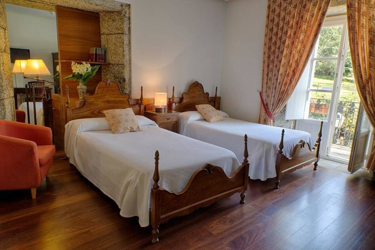 Una habitación de hotel con dos camas de madera, un sillón rojo, una mesita de noche con lámpara y un ventanal con cortinas.