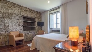 A bedroom with a stone wall, a bed, a chair, a TV, and a window with curtains. - Pazo de Sedor (Arzua)