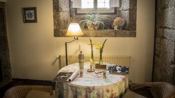 Una habitación acogedora con una mesa redonda, sillas de mimbre, una lámpara, un cuadro enmarcado y una ventana con plantas.