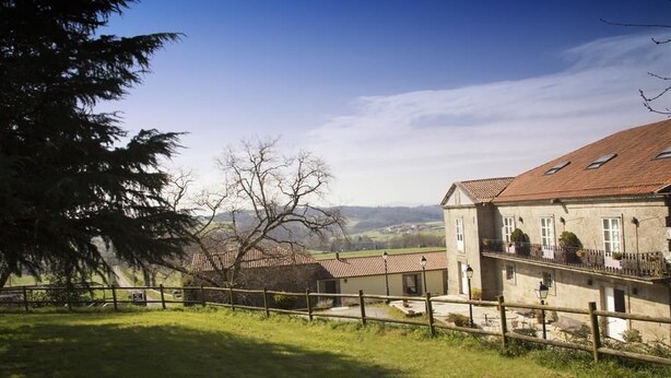 A rural setting with a house featuring a tiled roof, a wooden fence, and a view of distant hills. - Pazo de Sedor (Arzua)