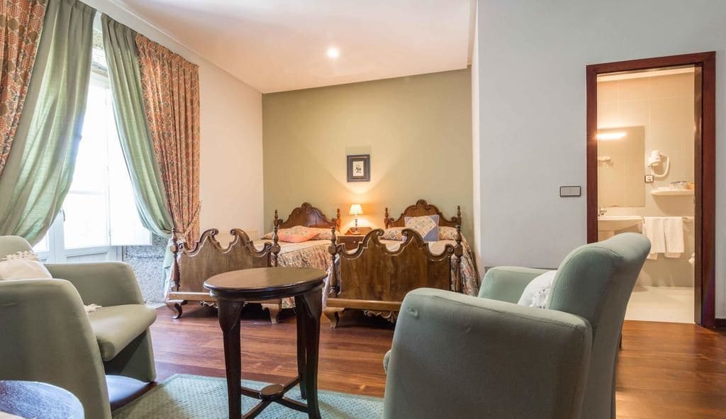 Habitación de hotel con cama, dos sillones, una mesita y un baño visible a través de una puerta abierta.