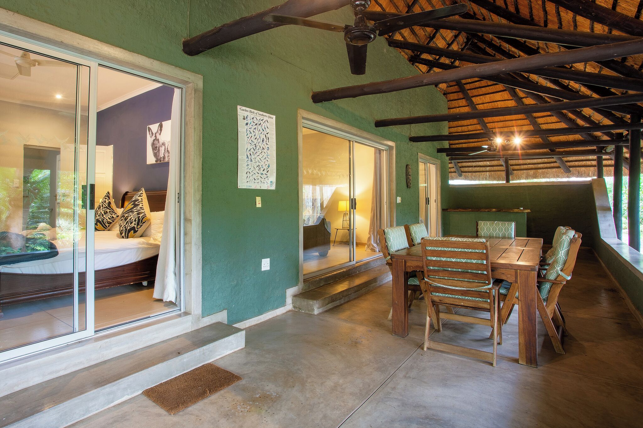 A dining area with a wooden table and chairs, a ceiling fan, and a view through glass doors to a bedroom.