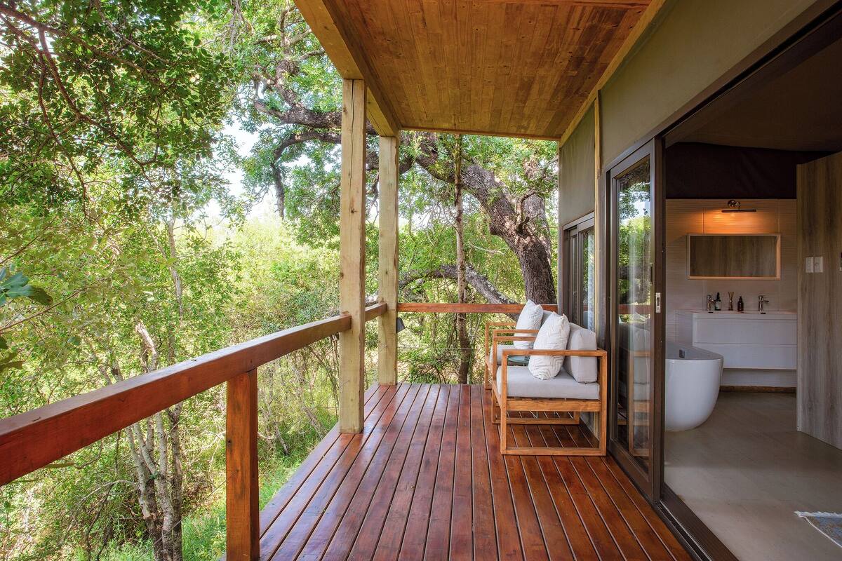 A wooden deck with a bench, a glass door leading to a bathroom, and a view of the surrounding forest.