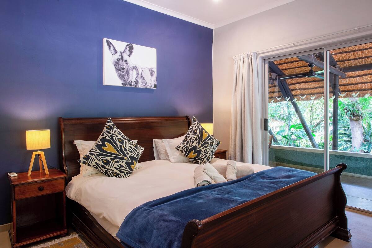 A bedroom with a bed, a nightstand, a window with a view of greenery, and a ceiling fan.