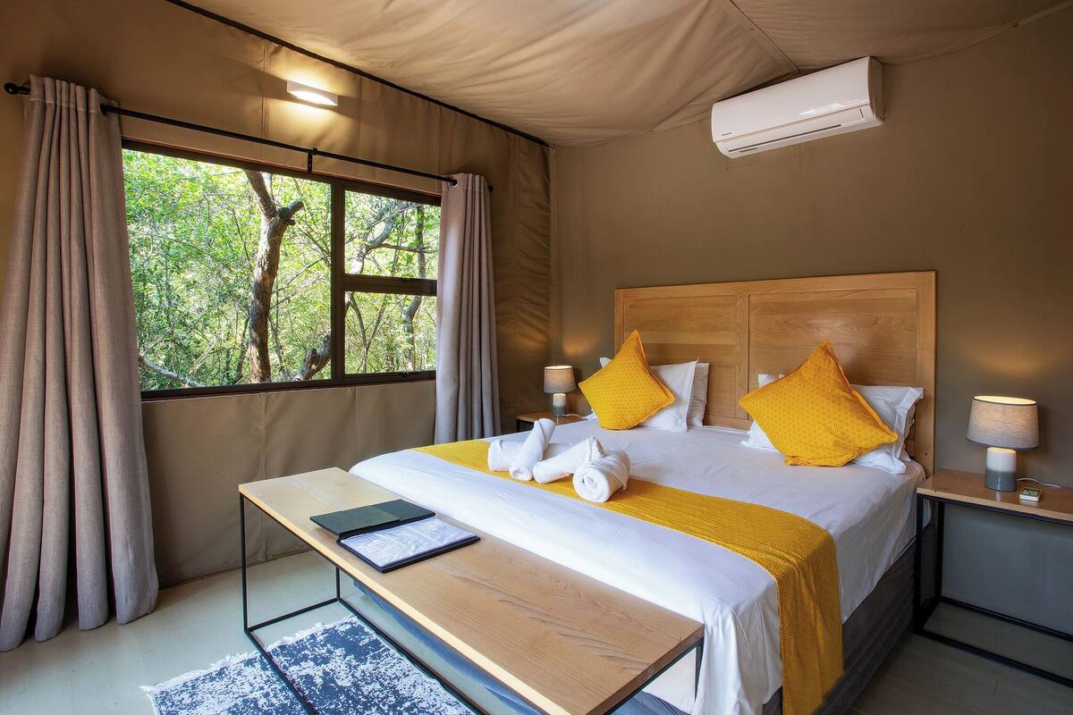 A bedroom with a bed, bedside tables, a desk, a window with curtains, and an air conditioning unit.