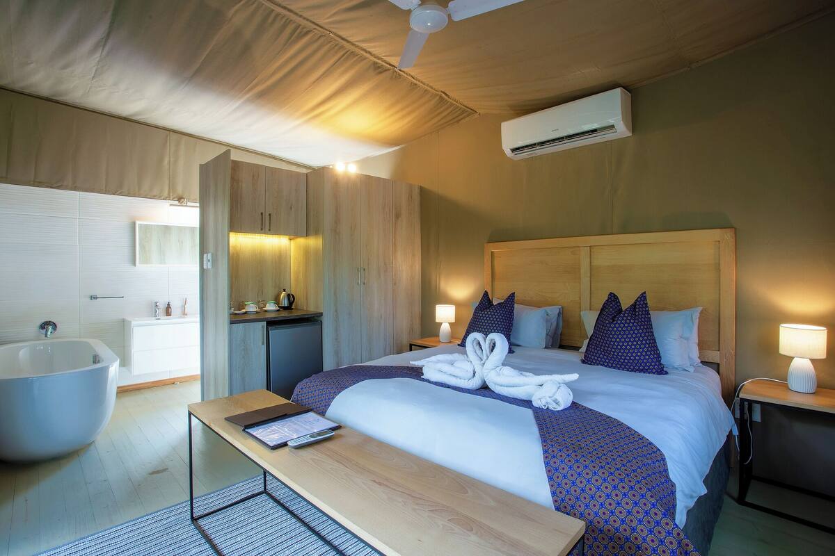 A hotel room with a large bed, a wooden headboard, a bedside table, a nightstand, a desk, a chair, a large bathtub, a ceiling fan, an air conditioning unit, and a mirror.