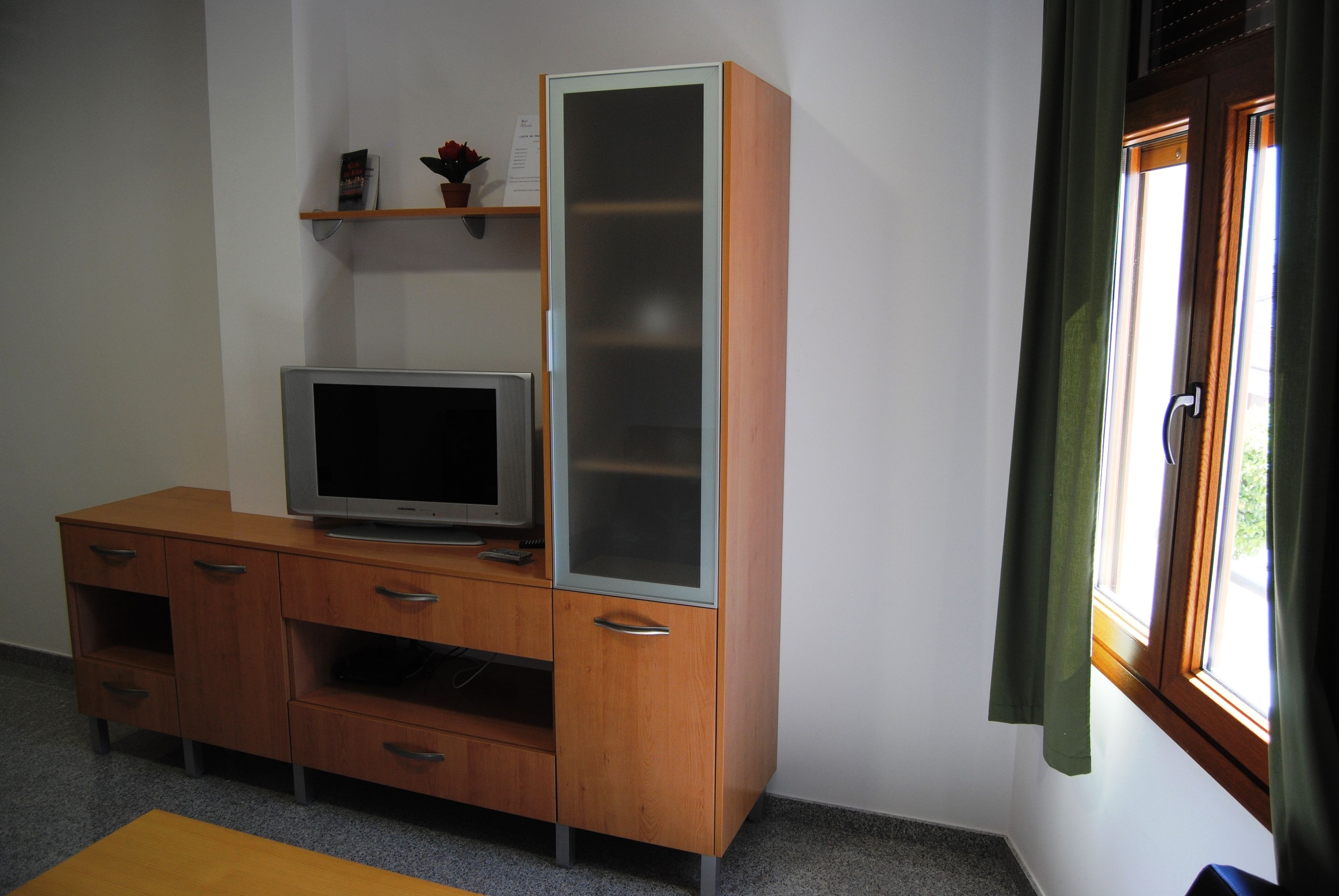 A room with a wooden TV stand, a flat-screen TV, and a window with curtains.