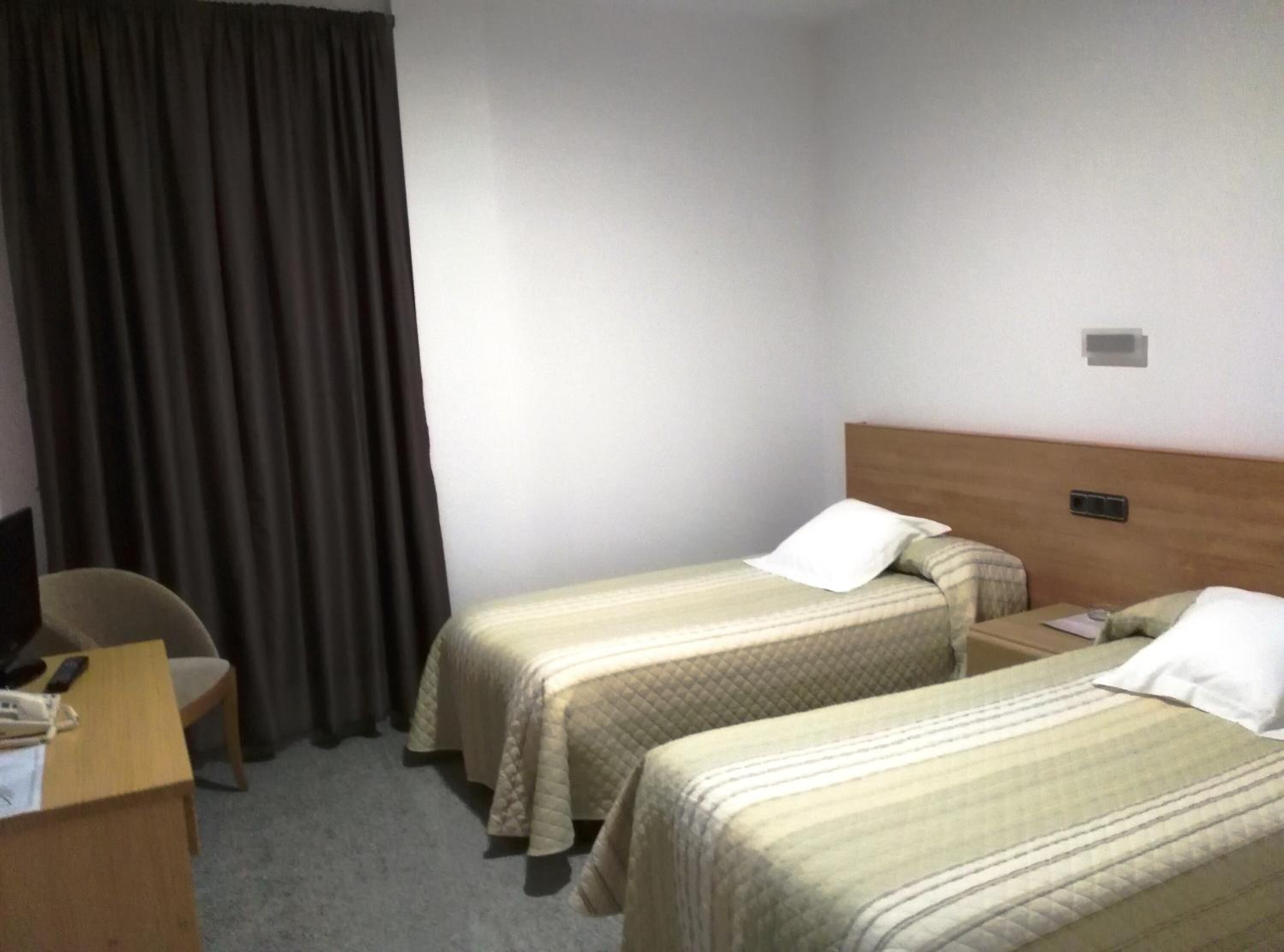 A hotel room with two single beds, a desk with a chair, and a large window with curtains.