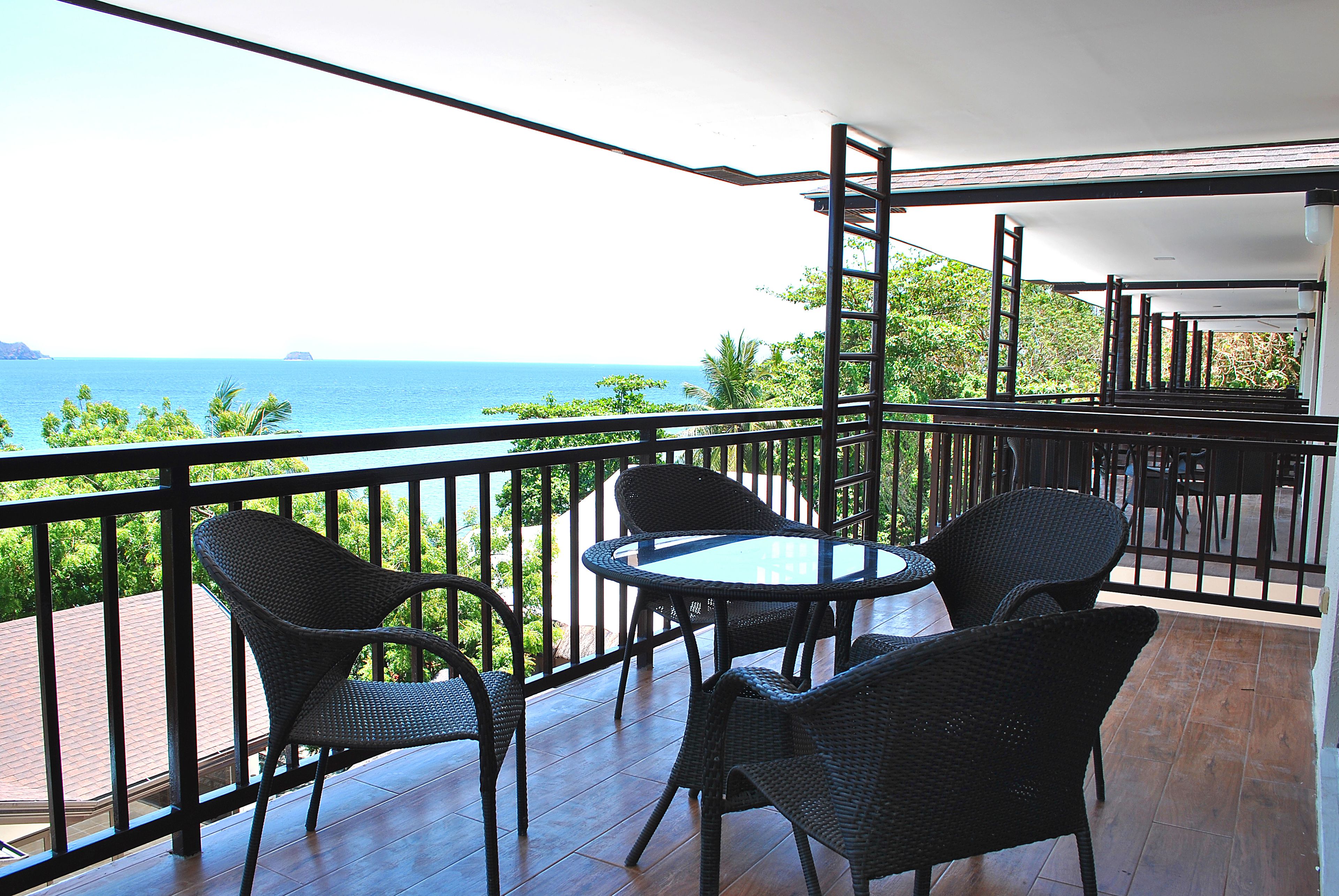 A balcony with wicker chairs and a glass table, overlooking a coastal view.
