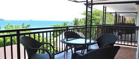 A balcony with wicker chairs and a glass table, overlooking a coastal view.