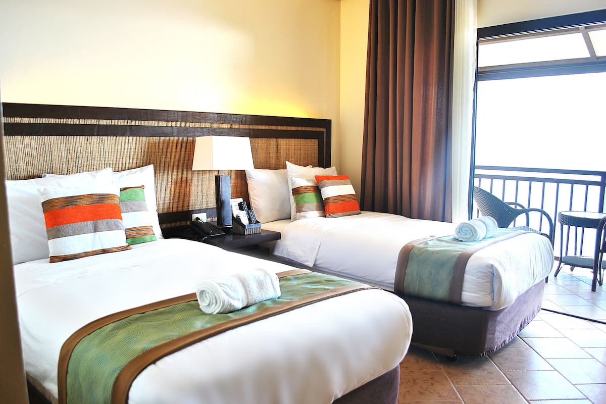 A hotel room with two beds, a nightstand with a lamp, and a view of the outside balcony.
