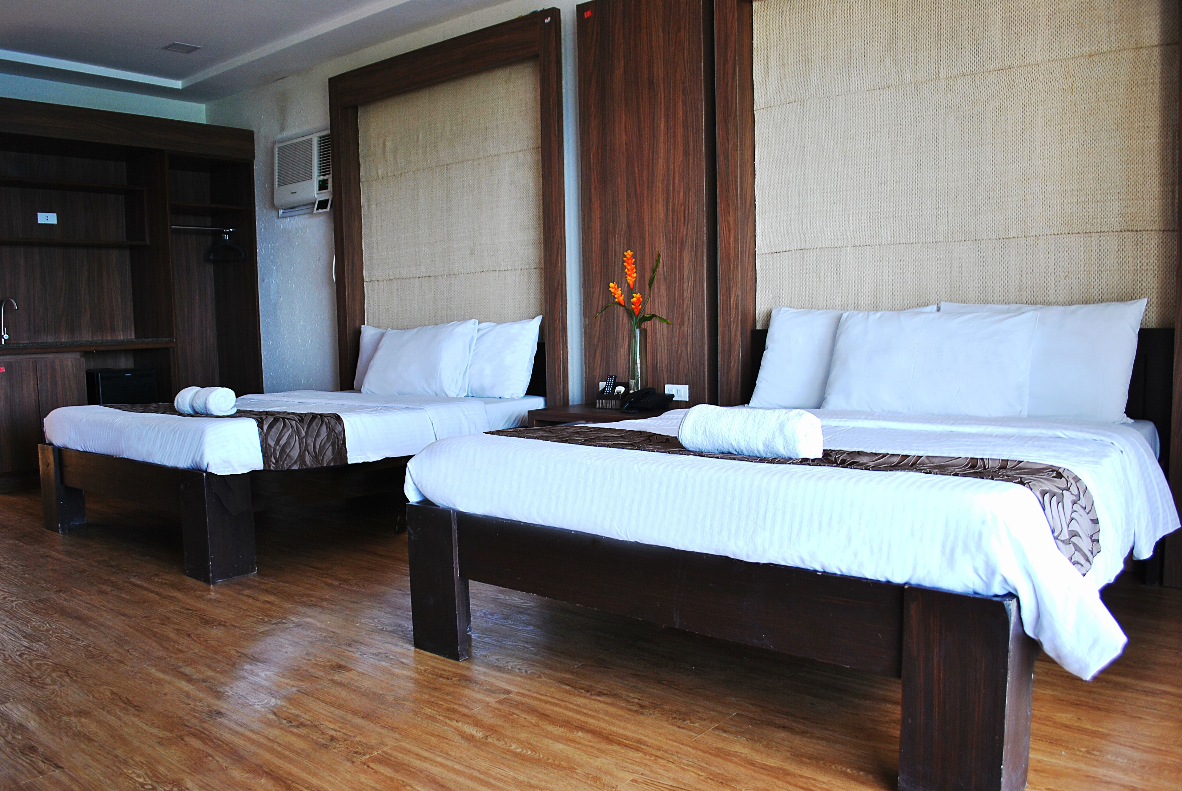Two single beds with wooden frames and white bedding, a small table with a vase of flowers, and a wardrobe in a hotel room.