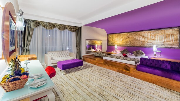 A modern living room with a purple accent wall, a white sofa, a purple ottoman, a wooden coffee table, and a large painting.