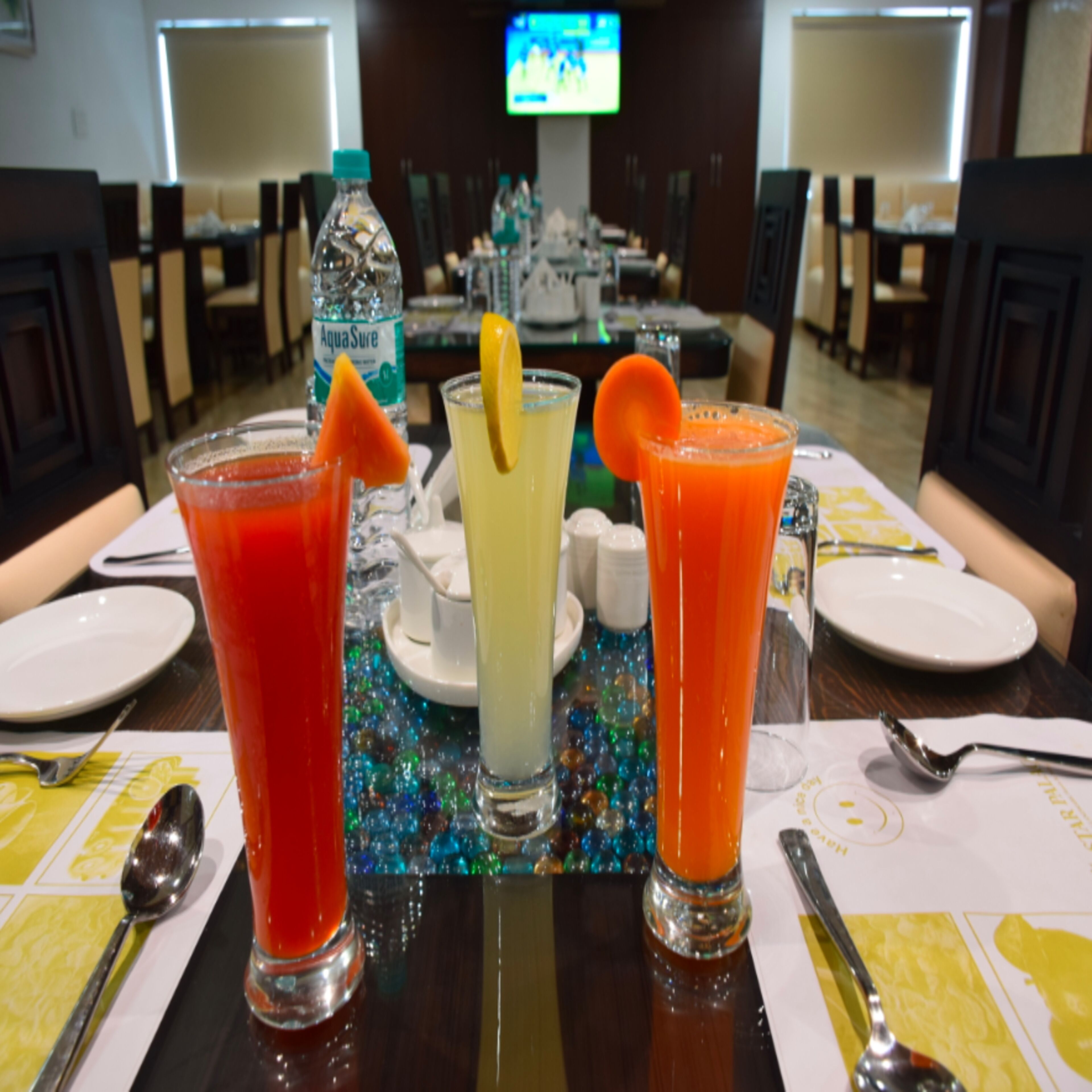 A table with three drinks, a bottle of water, and a television in the background.