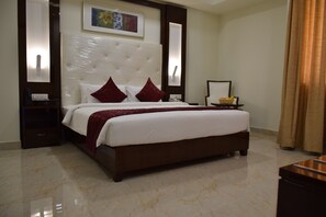 A large bed with a tufted headboard, two red pillows, a white bedspread, and a red throw blanket. There is a nightstand with a phone and a small table with fruit. - Star Palace Hotel (Rameshwaram)