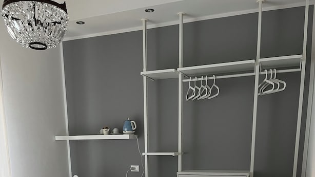 A bedroom with a tufted headboard, a bed with white linens, a bedside table with a lamp, a hanging light fixture, and a built-in wardrobe with shelves and hangers. - Fleur Blanche (Verona)