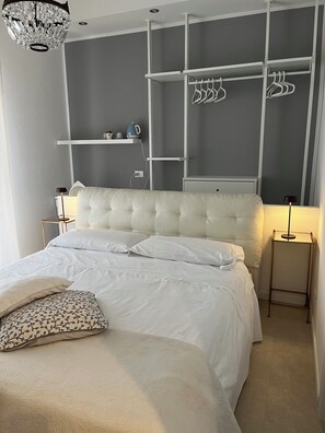 A bedroom with a tufted headboard, a bed with white linens, a bedside table with a lamp, a hanging light fixture, and a built-in wardrobe with shelves and hangers. - Fleur Blanche (Verona)