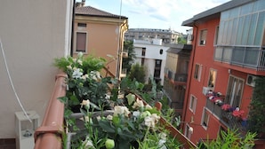 A narrow urban street with terracotta railings, a small garden with white flowers, and buildings with balconies. - Fleur Blanche (Verona)