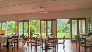 A dining area with multiple tables and chairs, large windows with a view of a lush garden. - Kitulgala Heritage Rest House (Kithulgala)