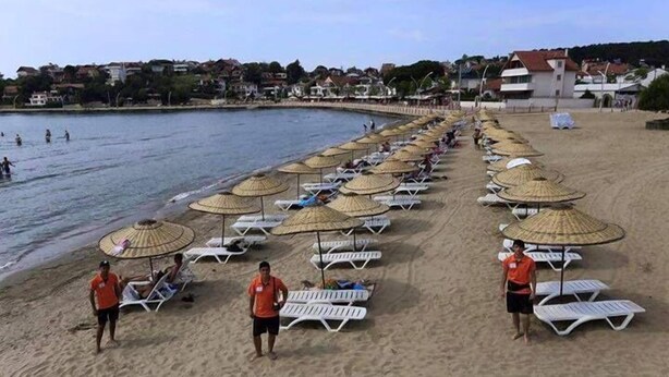 Een strand met rijen ligbedden en parasols, een paar mensen die wandelen, en huizen op de achtergrond.