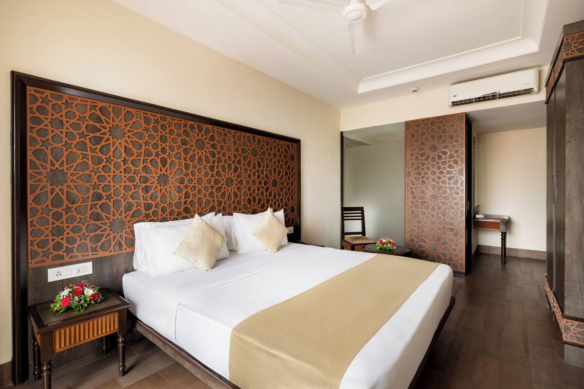A hotel room with a large bed, wooden headboard, bedside tables, a mirror, and a small table with a vase of flowers.