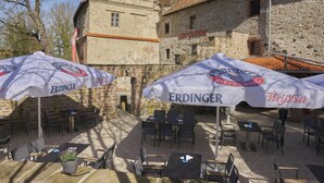 Un área de estar al aire libre con mesas y sillas bajo grandes sombrillas, un edificio de piedra al fondo y un letrero que dice "Weissbier".