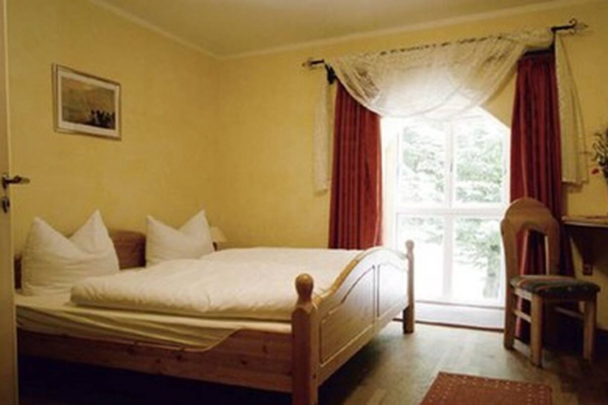 Een slaapkamer met een houten bed, een klein krukje, een bureau en een raam met gordijnen.