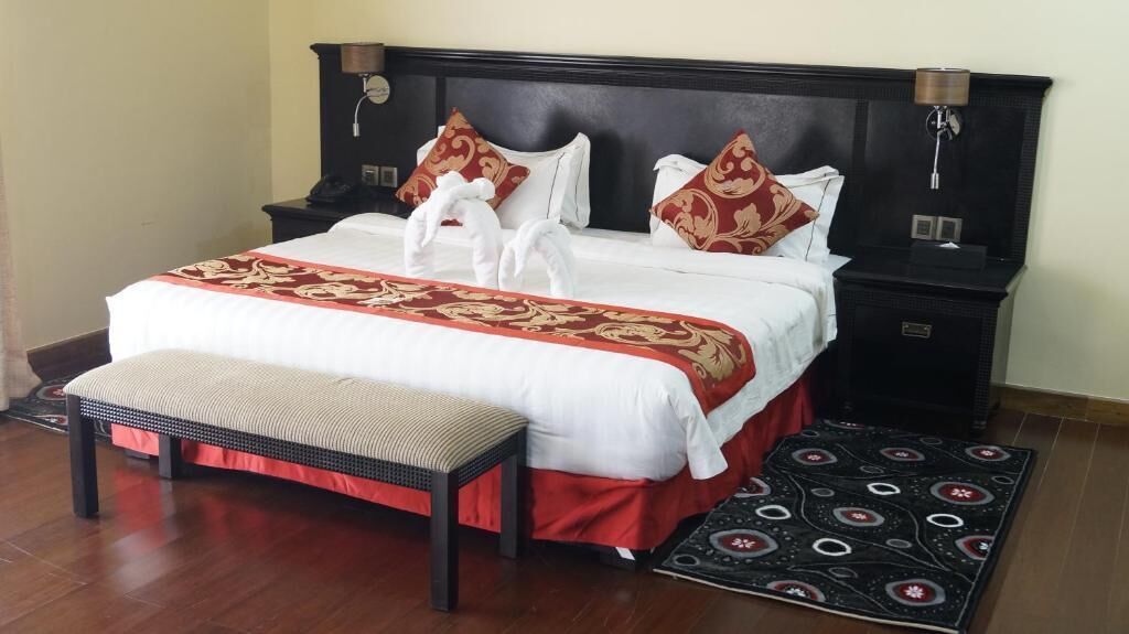 A neatly made bed with a red and white bedspread, two pillows, a bedside table with a lamp, and a bench at the foot of the bed.