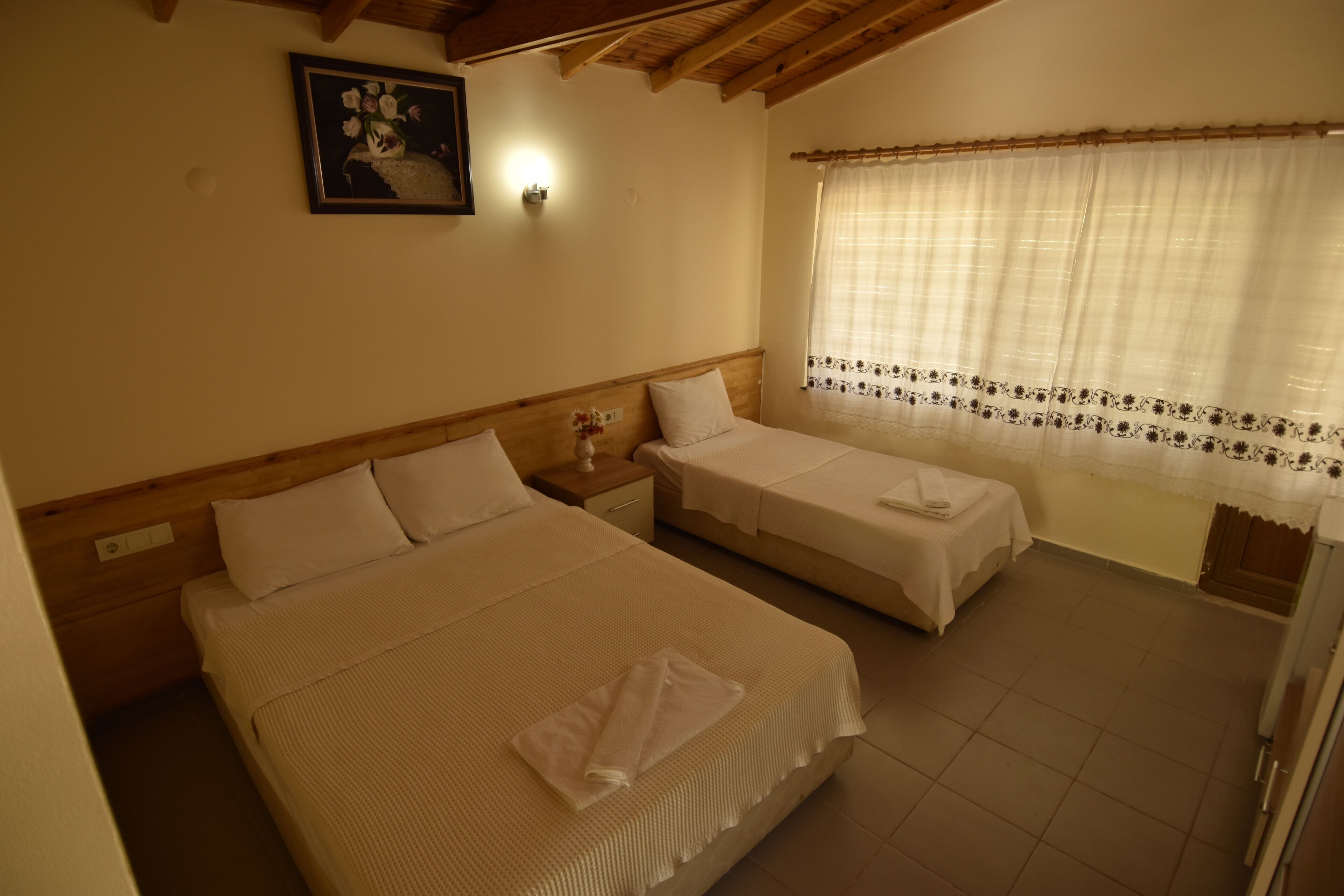 A room with two beds, a nightstand, a wall-mounted light, a window with curtains, and a framed picture on the wall.