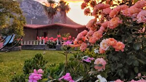 A garden with pink and white flowers, a building with a red roof, and mountains in the background. - Likya Adrasan (Kumluca)
