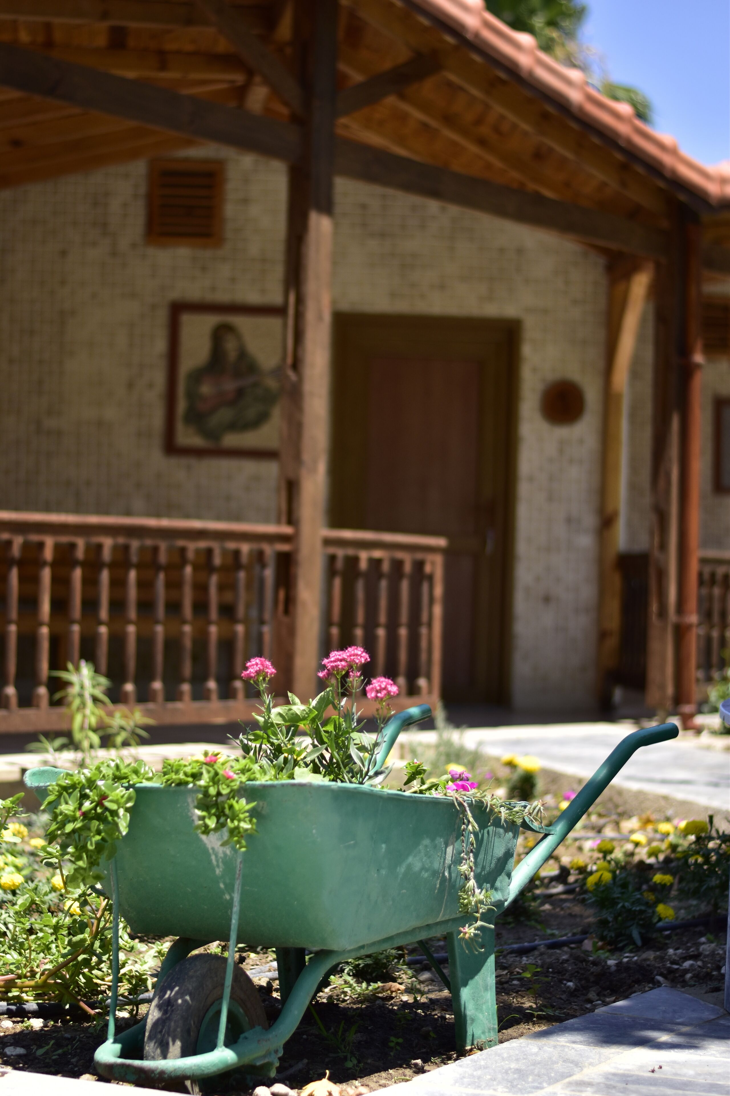 A green wheelbarrow with flowers in front of a rustic building with a tiled wall and a wooden balcony.