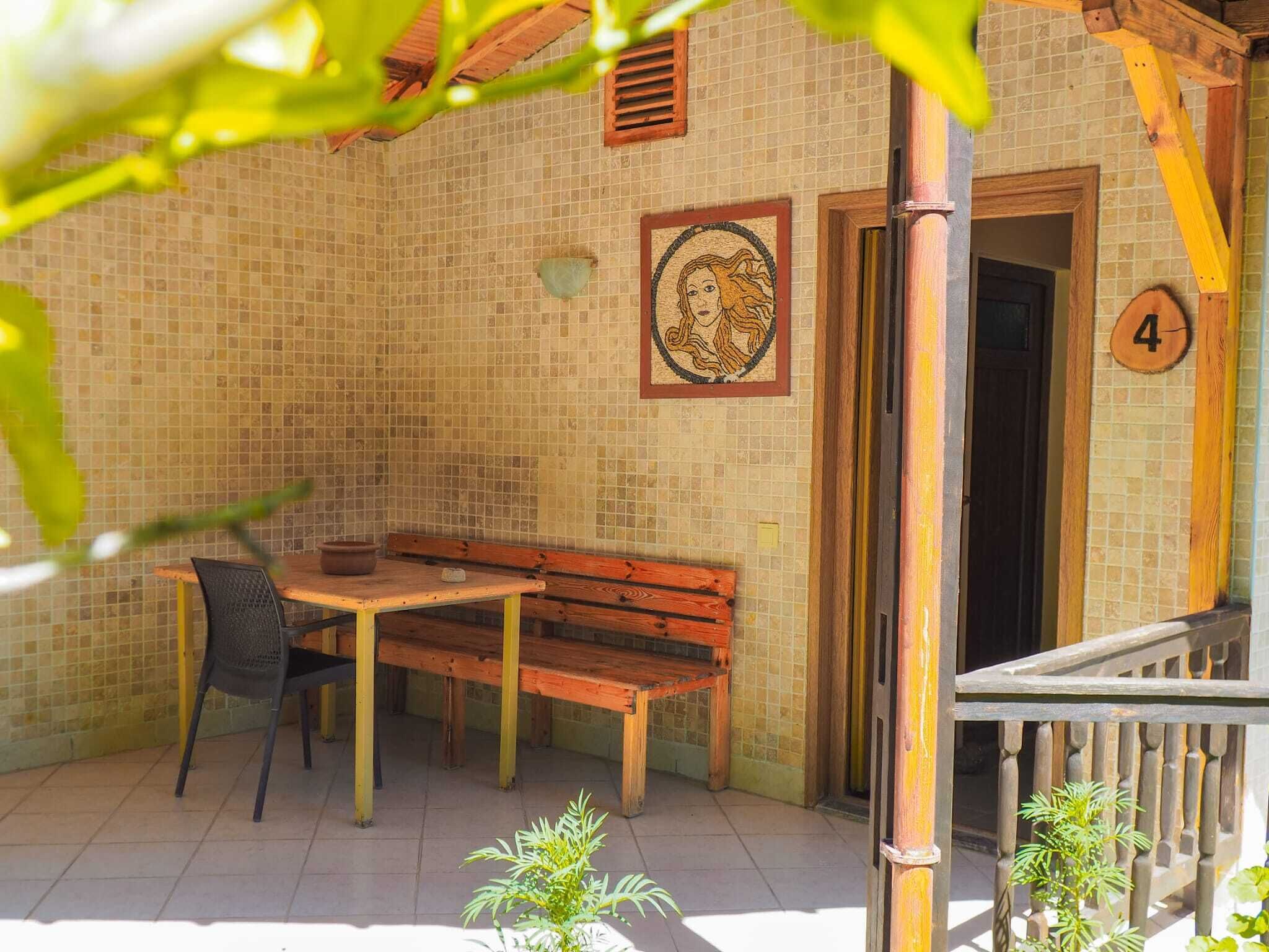 A small outdoor seating area with a wooden bench and table, a tiled wall with a mosaic, and a door marked with the number 4.