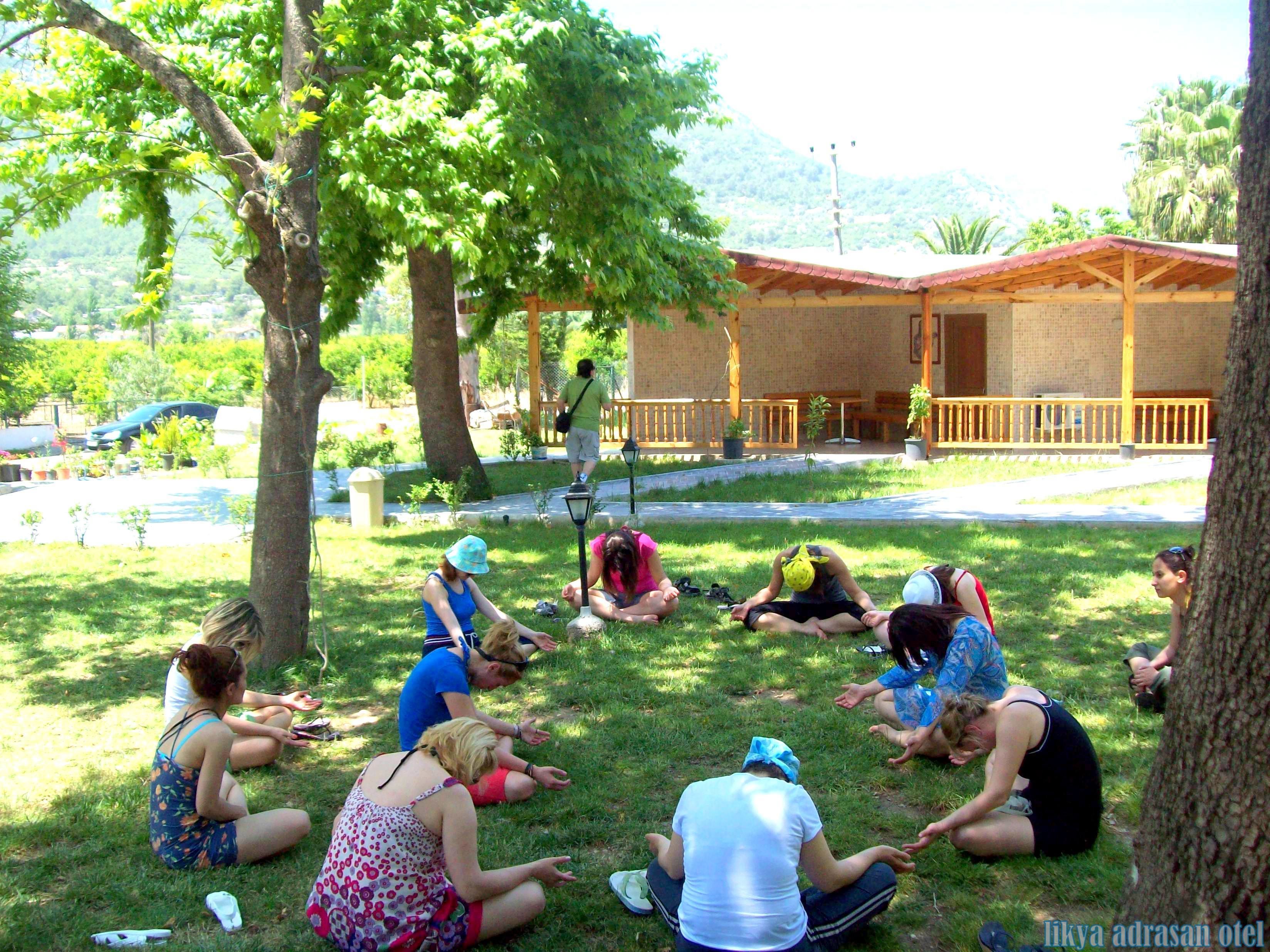 A group of people sitting on grass in a park-like setting with trees and a building in the background.