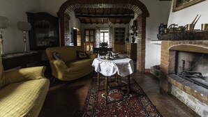 Een knus vertrek met een bank, een tafel met kantkleed, een patroonrijk vloerkleed en een open haard.