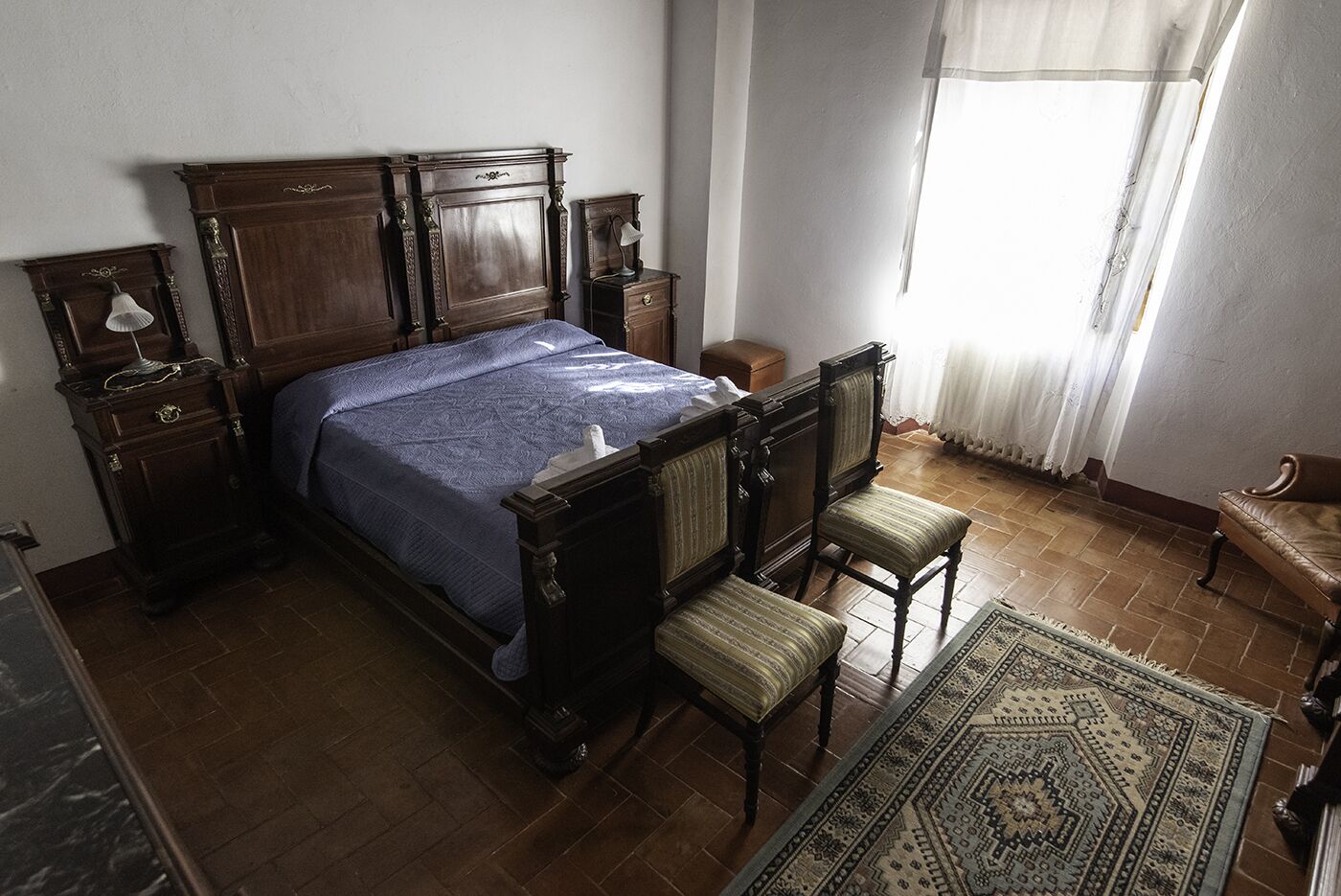 A traditional bedroom with a wooden bed, two bedside tables, a chair, a small table, and a window with curtains.