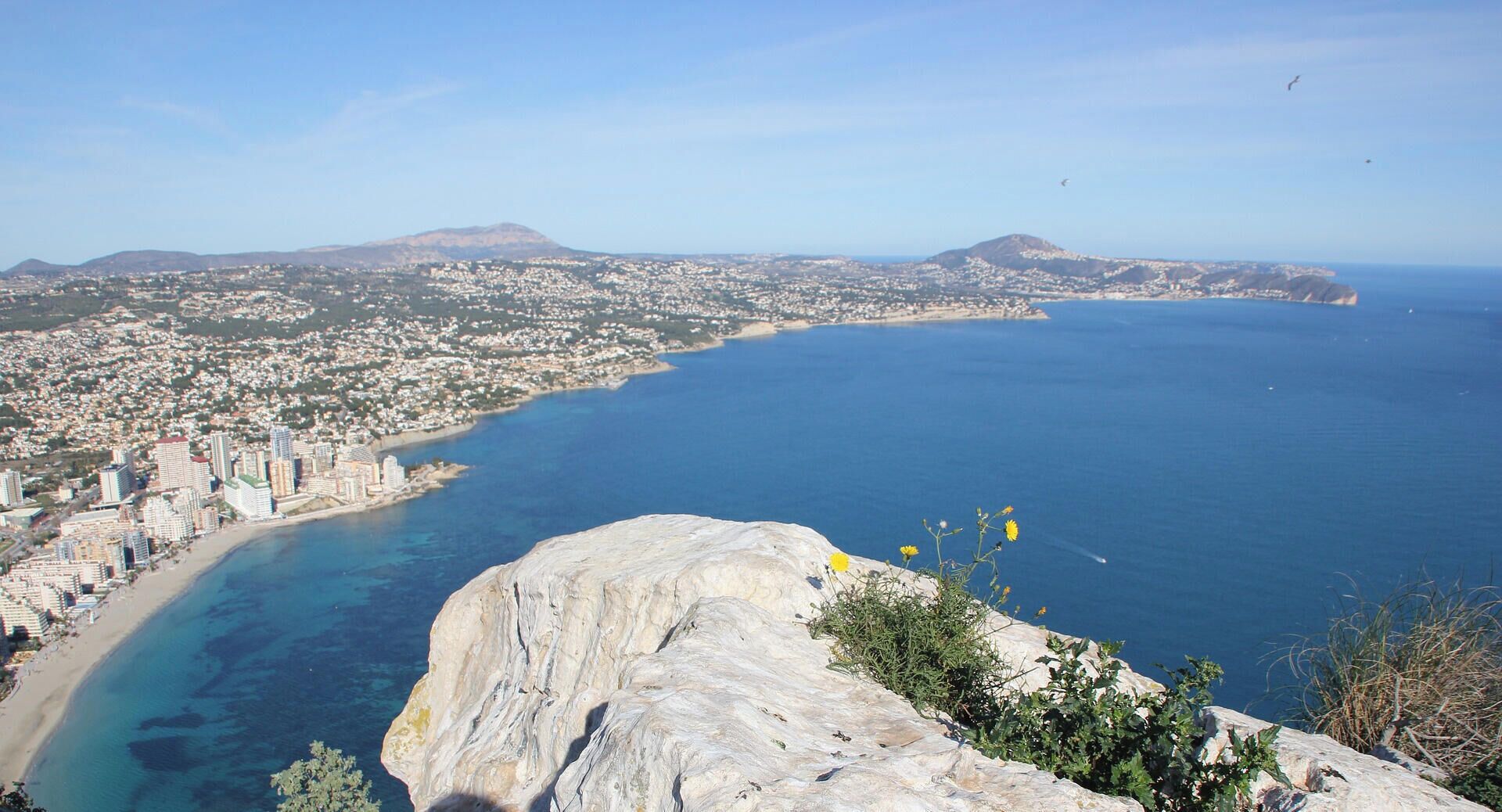 A coastal cityscape with a prominent rocky outcrop in the foreground, a clear blue sea, and a densely built urban area in the background.