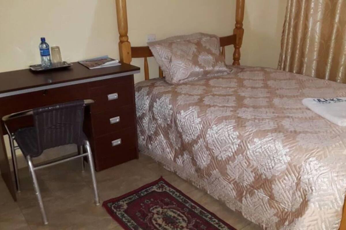 A bedroom with a wooden bed, a desk with a chair, a water bottle, and a small table.