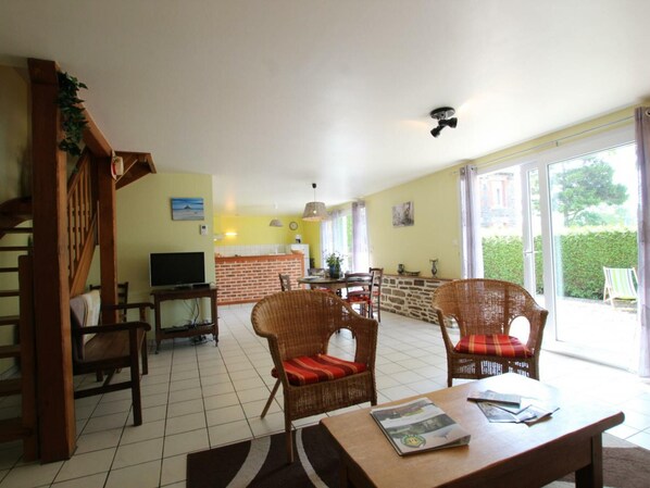 A spacious living area with a TV, wicker chairs, a dining table, and a kitchen in the background.
