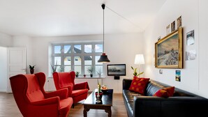 A living room with a red armchair, a black sofa, a wooden coffee table, and a large window with a view of the outside.