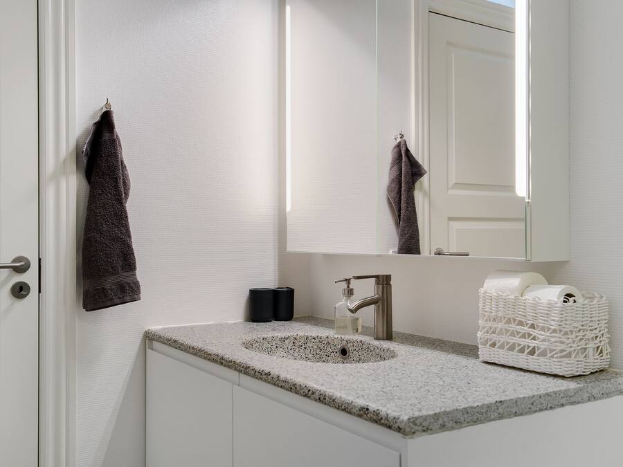 Un baño con encimera de granito, un lavabo, un espejo y una cesta de mimbre blanca sobre la encimera.