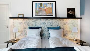 Una cama doble con cabecera azul y blanca estampada, dos mesitas de noche con lámparas, y una obra enmarcada sobre la cama.