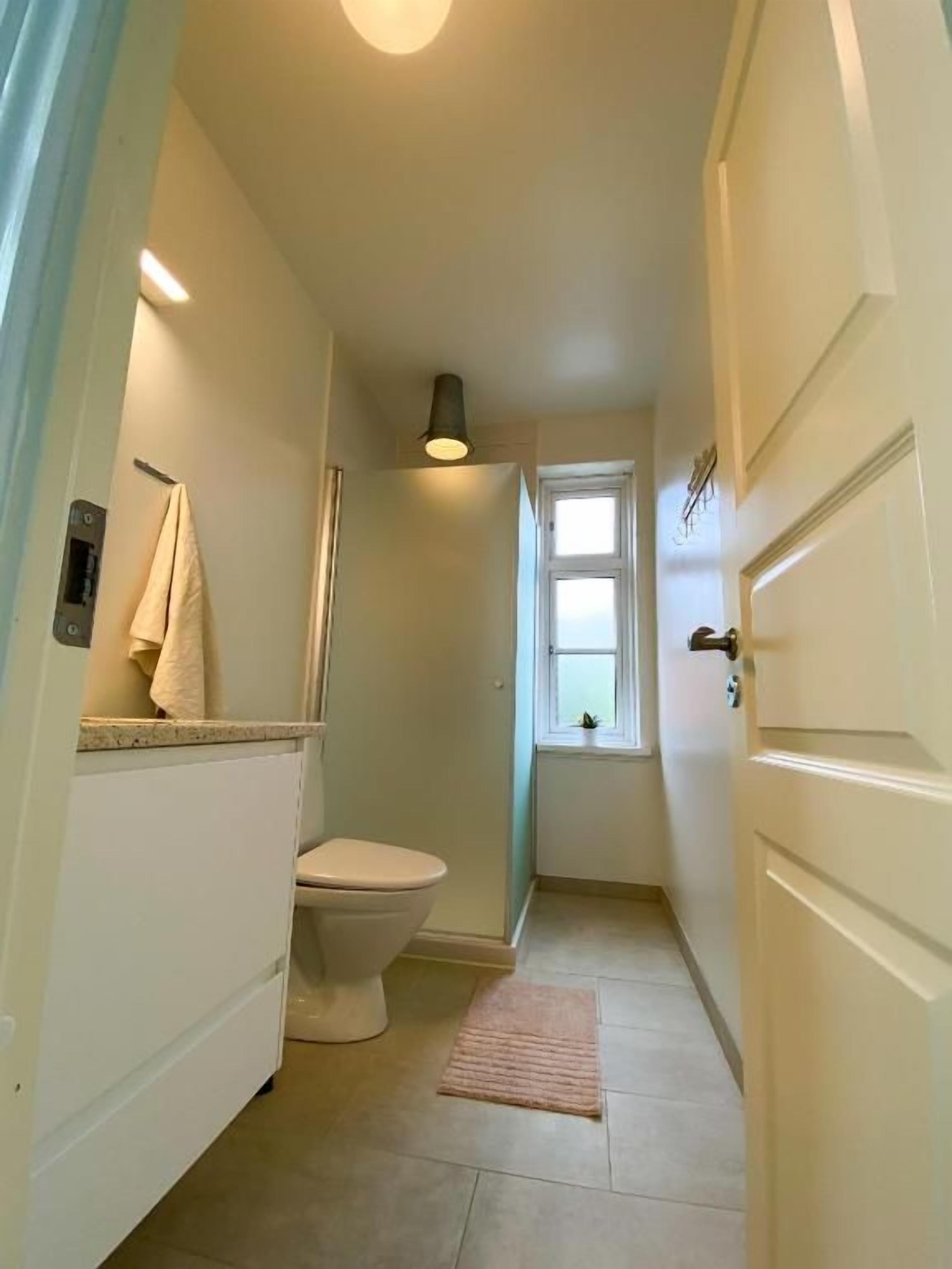 A narrow hallway leading to a bathroom with a toilet, a towel rack, and a window.