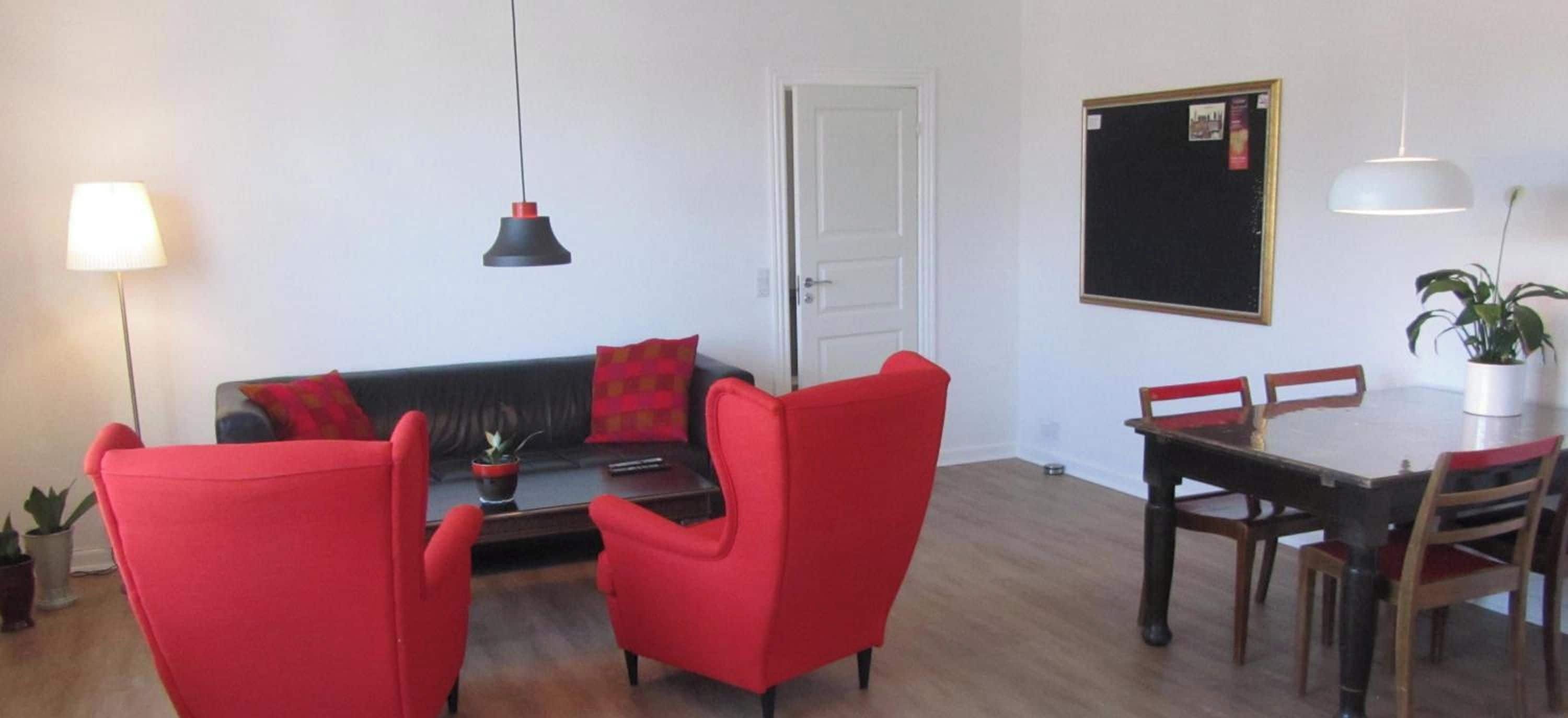 A living room with a sofa, two red chairs, a dining table with chairs, a lamp, a door, and a wall-mounted item.